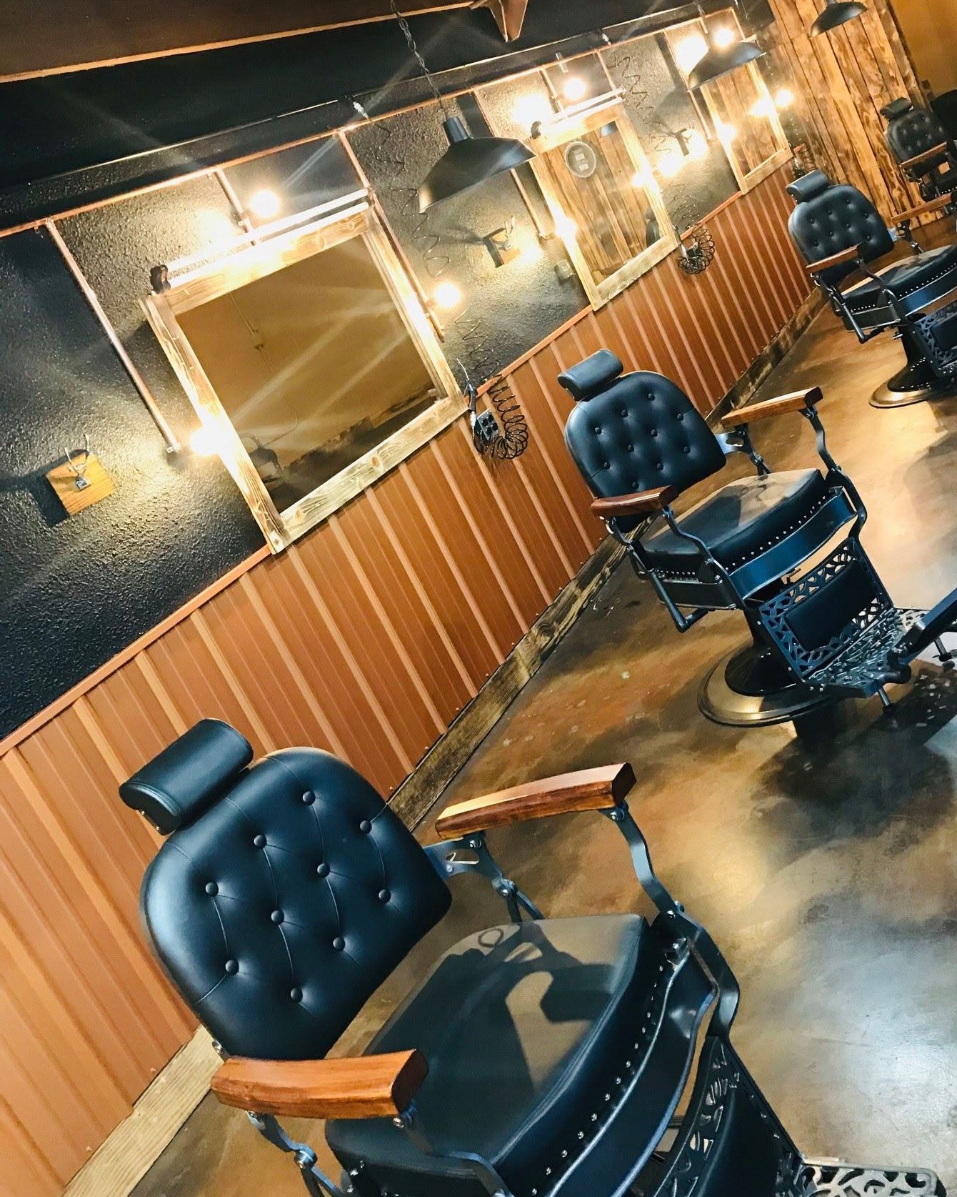 Inside Patrón Barbershop: a row of black tufted barber chairs along a copper-colored corrugated-metal wainscot, with dark textured walls, wood-framed lit mirrors and hanging lights above